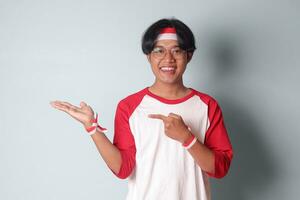 Portrait of attractive Asian man in t-shirt with red and white ribbon on head, pointing at something with finger. Isolated image on gray background photo