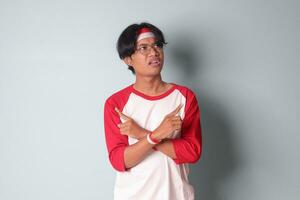 Portrait of attractive Asian man in t-shirt with red and white ribbon on head, choosing between 2 different options, holding two hands with empty space. Decision concept. Isolated image on gray photo