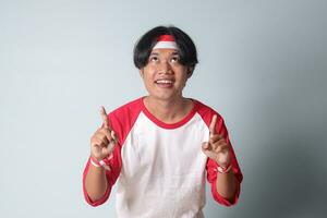 Portrait of attractive Asian man in t-shirt with red and white ribbon on head, pointing at something with finger. Isolated image on gray background photo