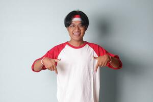 Portrait of attractive Asian man in t-shirt with red and white ribbon on head, pointing at something down with finger. Isolated image on gray background photo