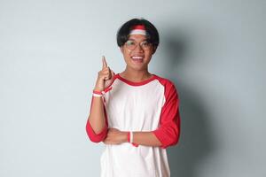 Portrait of attractive Asian man in t-shirt with red and white ribbon on head, presenting index finger and reminding of something to do. Isolated image on gray background photo