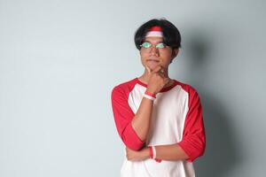 Portrait of attractive Asian man in t-shirt with red and white ribbon on head, standing against gray background, thinking about question with hand on chin photo