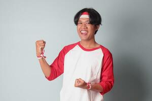 Portrait of attractive Asian man in t-shirt with red and white ribbon on head, raising his fist, celebrating Indonesia's independence day. Isolated image on gray background photo