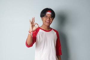 retrato de atractivo asiático hombre en camiseta con rojo y blanco cinta en cabeza, demostración Okay mano gesto y sonriente. aislado imagen en gris antecedentes foto