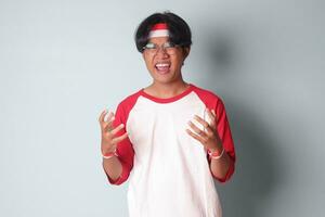 Portrait of attractive Asian man in t-shirt with red and white ribbon on head, shaking hands frustrated, feeling angry and mad at someone. Isolated image on gray background photo