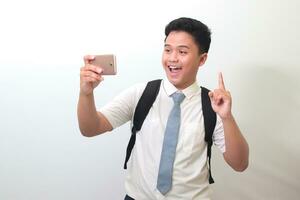 Indonesian senior high school student wearing white shirt uniform with gray tie explaining something with finger up while doing vlog on mobile phone. Isolated image on white background photo