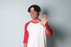 Portrait of attractive Asian man in t-shirt with red and white ribbon on head, counting five with fingers. Isolated image on gray background photo