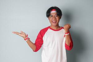 retrato de atractivo asiático hombre en camiseta con rojo y blanco cinta en cabeza, levantamiento su puño mientras demostrando producto. aislado imagen en gris antecedentes foto
