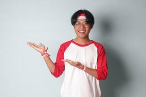 Portrait of attractive Asian man in t-shirt with red and white ribbon on head, pointing at something with finger. Isolated image on gray background photo