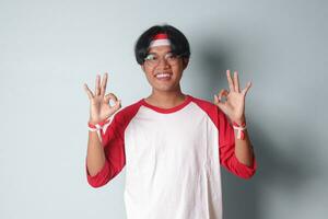 Portrait of attractive Asian man in t-shirt with red and white ribbon on head, showing ok hand gesture and smiling. Isolated image on gray background photo