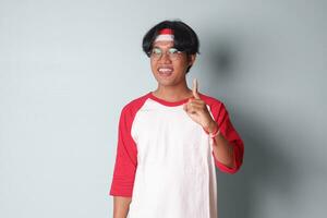 Portrait of attractive Asian man in t-shirt with red and white ribbon on head, presenting index finger and reminding of something to do. Isolated image on gray background photo