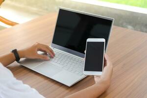 Close up of businessman hand touching blank screen of mobile phone with laptop beside on the table.  Display of smartphone and laptop mock up photo