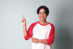 Portrait of attractive Asian man in t-shirt with red and white ribbon on head, pointing at something with finger. Isolated image on gray background photo