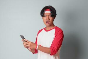 Portrait of shocked Asian man in t-shirt with red and white ribbon on head, holding mobile phone with surprised expression. Isolated image on gray background photo