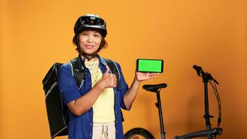 Courier holding mock up green screen smartphone, showing thumbs up hand gesturing. Delivery woman presenting chroma key touchscreen of phone in landscape mode, isolated over orange studio background photo