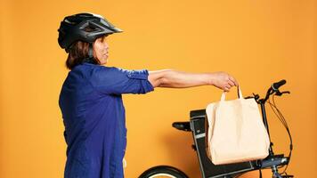 BIPOC food delivery worker reaching order address, waiting for customer to answer door. Courier woman greeting client, offering takeaway lunch bag, isolated over orange studio background photo