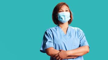 Close up shot of cheerful asian healthcare expert folding her arms, standing isolated over blue studio background. Happy BIPOC nurse wearing professional medical uniform, looking at camera photo