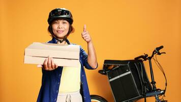 Portrait of happy courier looking at camera, showing thumbs up sign while delivering takeaway food. Smiling bike rider offering pizza boxes to client, isolated over orange studio background photo