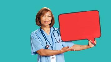 Joyful hospital employee holding red speech bubble sign of empty space for text. BIPOC experienced nurse presenting thought bubble cardboard, isolated over studio background, close up photo