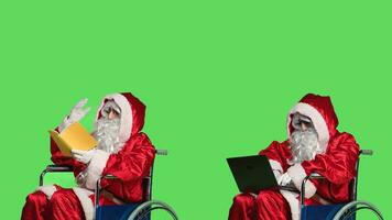 Saint nick with book and laptop sitting in wheelchair, dealing with impairment and reading novel story or browsing webpage. Young man dressed as father christmas against greenscreen. photo