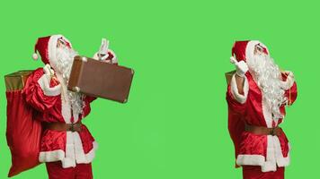 Santa hitchhiking with suitcase and sack full of gifts for children, making signs for transportation over greenscreen. Saint nick carrying briefcase and presents in iconic red bag. photo