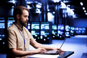 Computer scientist using laptop to patch up servers helping businesses manage databases, host websites and store files. Cloud computing business executive servicing supercomputers in data center photo