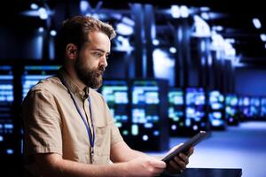 Skilled mechanic resolving performance related issues in server room. Expert utilizing tablet to identify operational problems causing high tech facility electronics slowdown photo