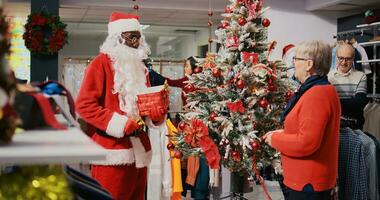 Employee in xmas adorn fashion boutique wearing Santa Claus costume and customers gathered around beautifully decorated Christmas tree in clothing store during winter holiday season photo