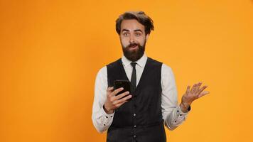 Restaurant employee messaging on duty and using phone at work, serving clients with meal orders. Classy waiter standing against yellow background in studio, checking smartphone social media apps. photo