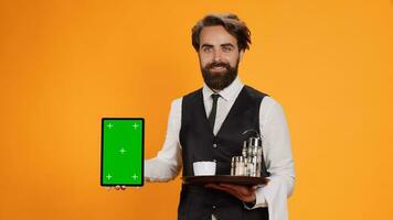 Friendly waiter shows tablet with greenscreen on camera, presenting blank copyspace mockup template. Experienced butler in suit holding device with isolated empty display and food tray. photo
