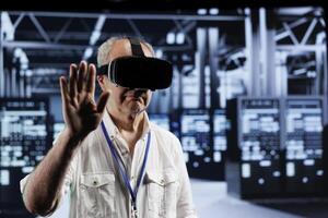 Senior skillful engineer immersed in virtual reality at data center, doing equipment maintenance. Expert using VR headset to optimize server system performance, harnessing cutting edge technology photo