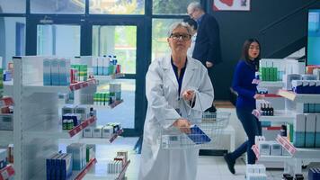 Elderly experienced pharmacologist in apothecary holding shopping basket, arranging merchandise on shelves. Senior pharmacist organizing pharmaceutical products in drugstore photo