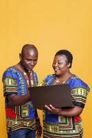 sonriente negro hombre y mujer compartiendo uno ordenador portátil para trabajo y en línea pasatiempo actividad. alegre africano americano Pareja vistiendo étnico ropa utilizando portátil computadora en estudio foto