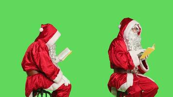 Wise santa claus reading book over greenscreen backdrop, enjoying poetry or literature book sitting on chair. Confident man dressed as father christmas with lecture hobby, knowledge. photo