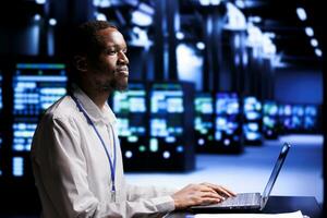 African american IT programmer in high tech facility using laptop to set up configuration management tools that enable automatic failover and load balancing, preventing server rigs system crash photo