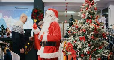 Joyful african american worker dressed as Santa Claus in Christmas ornate shopping mall clothing shop. Employee jingling xmas bells and greeting happy clients in festive adorn fashion store photo