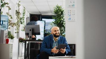 Company employee researching analytics on tablet, using past marketing statistics reports. Middle eastern man working with official documents to create small business development plan. photo