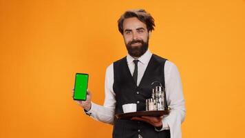 Young waiter shows greenscreen on camera, holding smartphone with blank copyspace display. Friendly elegant fine dining employee indicates isolated empty screen template in studio. photo