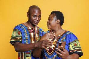 Smiling mid adult man and woman couple discussing and reading news on smartphone. Cheerful black wife and husband wearing ethnic clothes using mobile phone together and talking photo