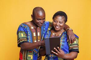 Smiling wife and husband with joyful expression hugging while using digital tablet together. Cheerful woman holding portable gadget while man tapping on touchscreen in studio photo