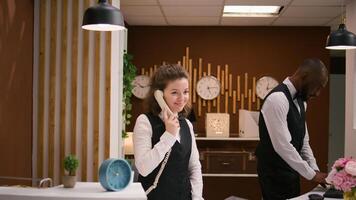 Hotel staff answers calls at reception, using landline phone to make remote bookings at front desk. Friendly receptionist keeping evidence of accommodations and room reservations. photo