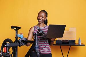 African American female inspecting and repairing bike with specialized tools and digital instructions from wireless computer. Active black woman examining and servicing broken bicycle with laptop. photo