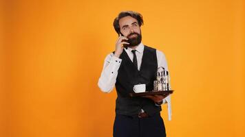 Restaurant waiter on smartphone call making reservations for fancy dinner, working in catering sector against yellow background. Man in formal attire talking to clients on remote phone line. photo