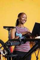 Focused young woman repairs broken bike, using specialized tools and searching for maintenance solutions on laptop. Vibrant african american female browsing on internet for new modern bicycle parts. photo