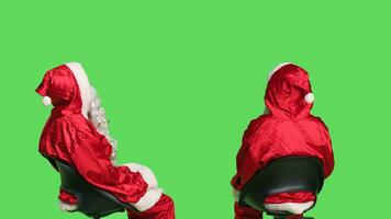 Santa claus posing on greenscreen background, young man dressed as saint nick sitting on chair and waiting. Acting impatient while he wears iconic festive costume with white beard. photo