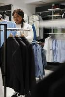 africano americano mujer seleccionando atuendo mientras hojeada mediante ropa bastidores en compras centrar Moda boutique. sonriente cliente elegir formal vestir para compra en Tienda foto