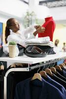 Jackets hanging on rack and accessories on shelf in clothing store while assistant dressing mannequin on blurred background. Male blazers and belts in department shopping mall photo