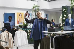 Young arab man internet influencer live streaming using smartphone in clothing store while showcasing jacket. Boutique client choosing outfit and showing apparel on hanger on mobile phone front camera photo