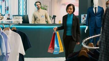 Female customer enjoying buying clothes from retail shop, doing dance moves in modern clothing store. Asian person feeling happy dancing in shopping center, holding shopping bags. photo