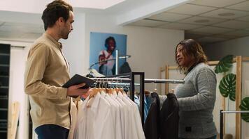 Boutique employee doing inventory near store aisles, checking stock and quantity of fashionable clothes holding tablet. Young man assistant looking at merchandise in mall shop. Handheld shot. photo